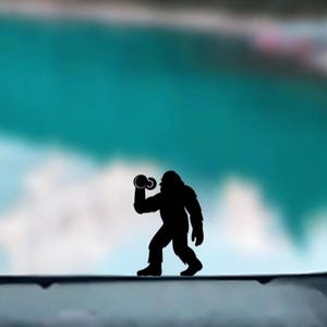 May include: A black silhouette of Bigfoot lifting weights, set against a blurred backdrop of a turquoise lake and green trees. The figure is positioned on a dark surface, creating a playful contrast.