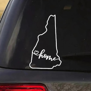 May include: White vinyl decal of the state of New Hampshire with the word "home" in a cursive font. A heart shape is incorporated into the design. The decal is affixed to the rear window of a vehicle.