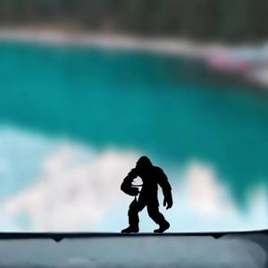 May include: Silhouette of a Bigfoot figure holding a camera, set against a blurred backdrop of a turquoise lake and green trees. The dark figure is walking on a light-colored surface, creating a contrast with the natural scenery.