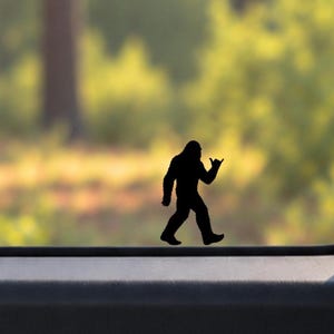 May include: A black vinyl decal of a silhouette of Bigfoot walking and making a shaka hand gesture. The decal is applied to a horizontal glass surface, showing the creature in profile with a hunched posture and long arms.
