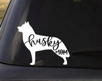 May include: A white vinyl decal applied to a car window, featuring the silhouette of a husky dog with the text "husky MOM" integrated into the body of the animal in a cursive script font. The design is solid white and stands out against the glass.