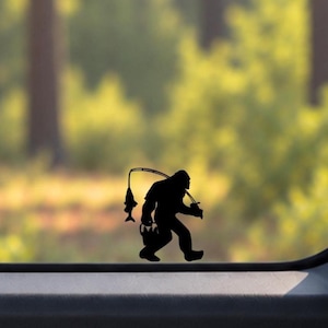 May include: A black vinyl decal sticker applied to a glass surface, featuring a silhouette of the creature known as Bigfoot. The figure is depicted walking while holding a fishing rod with a fish hanging from the line and carrying a bucket.