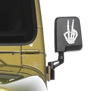 May include: A black rectangular vehicle side mirror featuring a white graphic decal of a skeleton hand making a peace sign. The mirror is attached to the olive green metal body of a vehicle, positioned near the front windshield and door hinge.