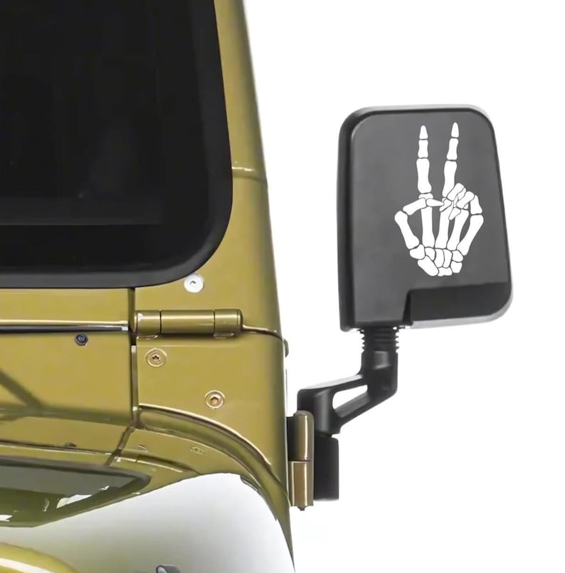 May include: A black rectangular vehicle side mirror featuring a white graphic decal of a skeleton hand making a peace sign. The mirror is attached to the olive green metal body of a vehicle, positioned near the front windshield and door hinge.
