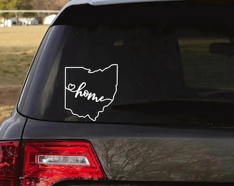 May include: A white vinyl decal applied to a dark vehicle rear window. The decal features the outline of the state of Ohio with the word home written in a cursive script font, incorporating a small heart shape into the letter h.