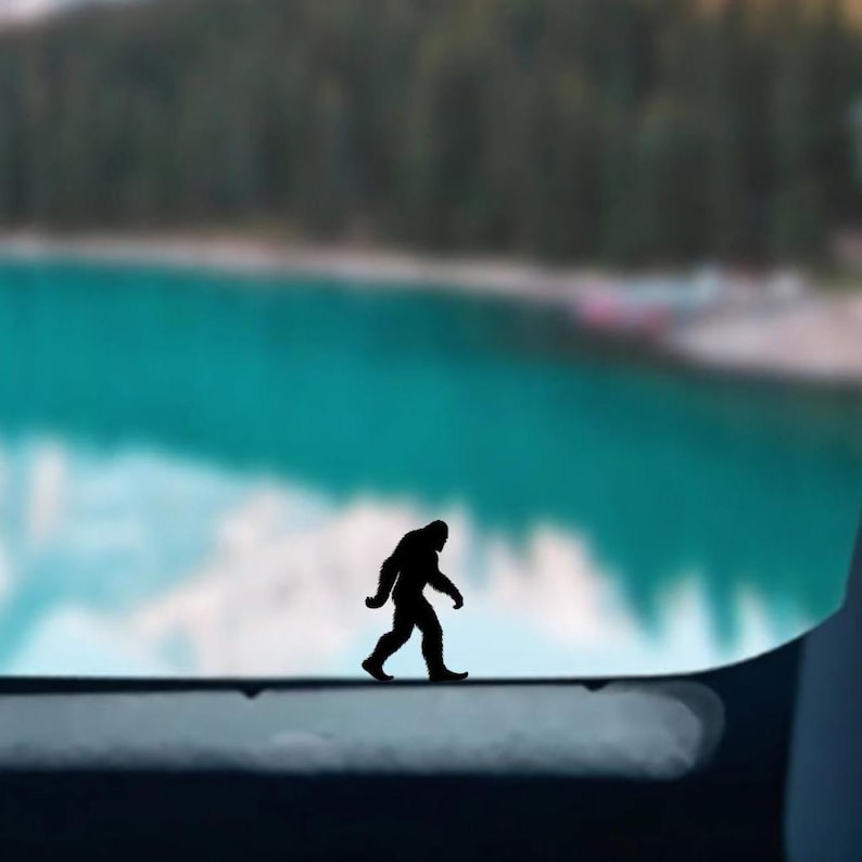 May include: Silhouette of Bigfoot walking across a blurry background of a lake and trees.