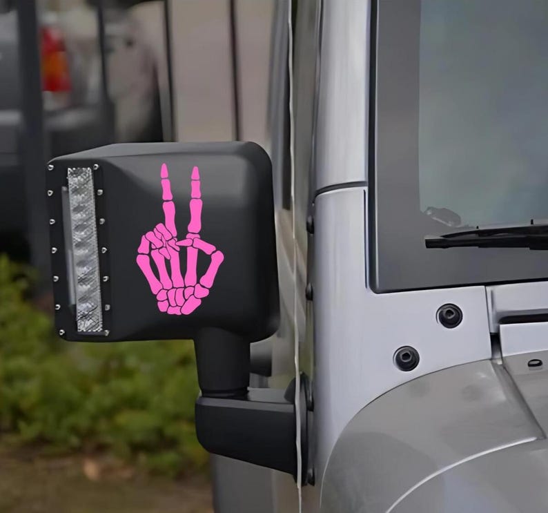 May include: A bright pink vinyl decal featuring a skeletal hand making a peace sign gesture, applied to the black plastic housing of a vehicle side mirror. The mirror assembly includes a vertical rectangular LED light strip with visible screws.