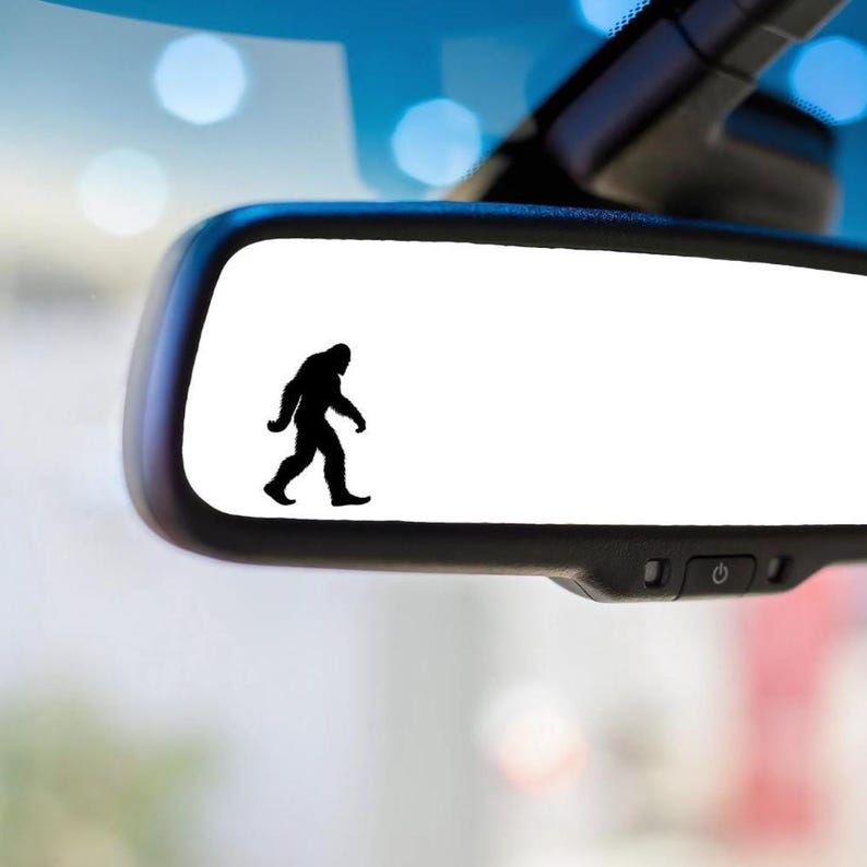 May include: A black silhouette of Bigfoot walking is printed on a white rearview mirror.