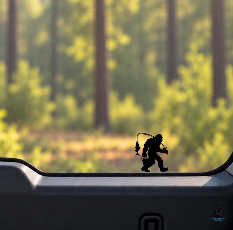 May include: A black vinyl decal sticker applied to a glass surface, featuring a silhouette of the creature known as Bigfoot. The figure is depicted walking while holding a fishing rod with a fish hanging from the line and carrying a bucket.