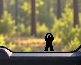 May include: A black vinyl decal featuring the silhouette of a Bigfoot creature standing upright and using its hands to form a heart shape above its head, applied to the surface of a vehicle window.