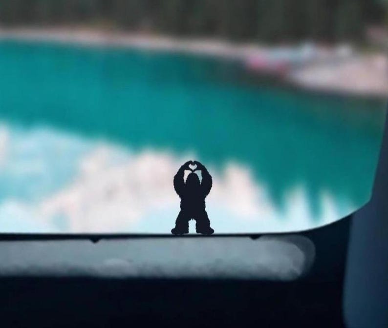 May include: A black silhouette of a bear making a heart shape with its paws, against a blurry background of a lake and trees.
