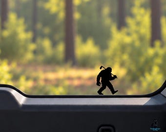 May include: A black vinyl decal of a silhouette of Bigfoot walking while carrying a golf club over a shoulder. The decal is applied to a glass surface, showing a clear, dark, solid figure against a blurred outdoor forest background.