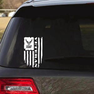 K&ouml;nnte beinhalten: Ein wei&szlig;er Vinyl-Aufkleber mit einer amerikanischen Flagge und den Worten "Veteran" und "USAF". Der Aufkleber befindet sich auf der Heckscheibe eines schwarzen Autos.