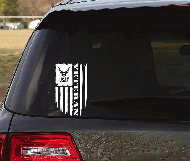 K&ouml;nnte beinhalten: Ein wei&szlig;er Vinyl-Aufkleber mit einer amerikanischen Flagge und den Worten "Veteran" und "USAF". Der Aufkleber befindet sich auf der Heckscheibe eines schwarzen Autos.