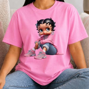 May include: A light pink short-sleeved t-shirt featuring a graphic print of a cartoon character with black hair, large eyes, and red lips. The character wears a pink long-sleeved top, blue jeans, and pink sneakers while sitting in a relaxed pose.