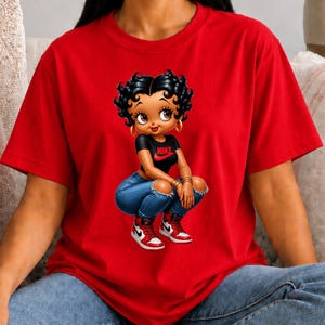 May include: A bright red short-sleeved t-shirt featuring a graphic of a cartoon character with black hair, wearing a black t-shirt with a red Nike logo, blue distressed jeans, and red and white high-top sneakers, crouching in a central position.