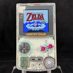 以下が含まれることがあります： 透明なゲームボーイカラー携帯ゲーム機。画面には「ゼルダの伝説 夢をみる島DX」のタイトル画面が表示されています。本体には黒い十字キー、2つのマゼンタ色のA・Bボタン、銀色のスタート・セレクトボタンが配置されています。