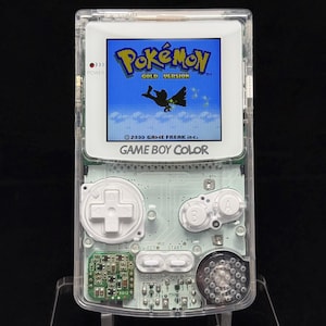 Può includere: Una console di gioco portatile Game Boy Color trasparente con un guscio in plastica chiara che rivela i circuiti interni. Lo schermo mostra la schermata del titolo di Pokémon Gold Version con una silhouette di Ho-Oh e il testo 2000 GAME FREAK inc.