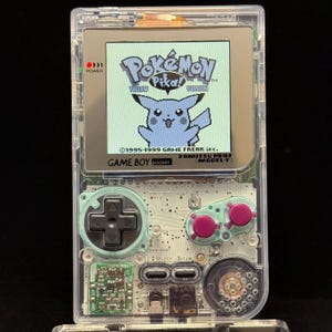 May include: A clear Game Boy Pocket handheld console featuring a screen displaying the Pokémon Yellow Version title with a Pikachu graphic. The device includes a black directional pad, two magenta buttons, and internal circuit board components visible.