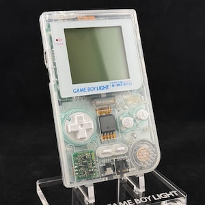 May include: A clear, transparent handheld video game console with a visible internal circuit board. The device features a square screen, a white directional pad, A and B buttons, and text reading GAME BOY LIGHT FAMITSU 500 MODEL-F02.