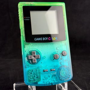 Op de afbeelding: Een draagbare Game Boy Color spelcomputer met een doorzichtige behuizing in een levendig kleurverloop dat van limoengroen aan de bovenkant naar helder cyaan aan de onderkant loopt. Het bevat een zwart schermframe, grijze knoppen en een luidspreker.
