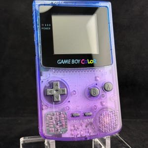 Pode incluir: Uma consola de jogos portátil Game Boy Color com uma estrutura de plástico roxo translúcido. O dispositivo inclui uma moldura de ecrã preta com o texto GAME BOY COLOR, um pad direcional cinzento, botões A e B cinzentos e botões Select e Start cinzentos.