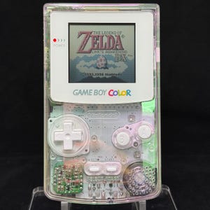 以下が含まれることがあります： 内部回路が見える透明なシェルを備えた、透明なゲームボーイカラー携帯ゲーム機。画面には「ゼルダの伝説 夢をみる島DX」のタイトル画面が表示されています。白い十字キーとAボタン、Bボタンが特徴です。