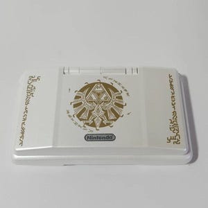 Könnte beinhalten: Eine weiße tragbare Spielkonsole mit einem goldenen Triforce-Emblem in der Mitte und goldener Hylian-Schrift auf der linken und rechten Seite. Das Wort Nintendo ist in Schwarz unten in der Mitte des Geräts aufgedruckt.