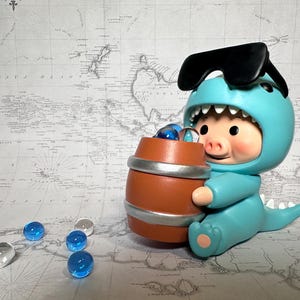 May include: A blue and white toy pig wearing sunglasses and a dinosaur costume holds a brown barrel filled with blue marbles. The pig is sitting on a white and blue map.