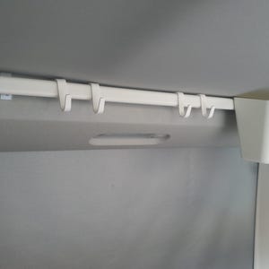 Könnte beinhalten: Ein weißes, an der Decke montiertes Aufbewahrungssystem mit einer horizontalen Stange und mehreren Haken. Ein weißer, rechteckiger Behälter ist am Ende befestigt. Das System dient zum Organisieren von Gegenständen in einem Fahrzeug.