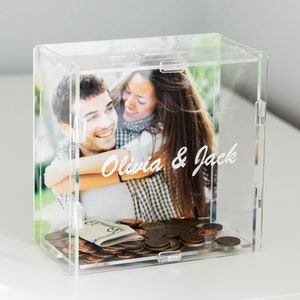May include: Clear acrylic box with a photo of a couple inside. The photo shows a man and a woman smiling and hugging. The text "Olivia & Jack" is written on the photo. The box is filled with coins.
