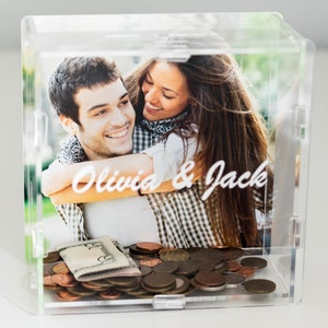 May include: Clear acrylic money box with a printed photo of a smiling couple embracing. The photo is labeled "Olivia & Jack" in white script. The box contains various coins and paper currency.