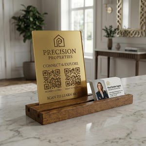 Op de afbeelding: Een goudkleurig rechthoekig bord met de tekst PRECISION PROPERTIES, CONNECT & EXPLORE en SCAN TO LEARN MORE, voorzien van twee QR-codes. Het bord staat in een bruine houten standaard naast een visitekaartje voor een makelaar.