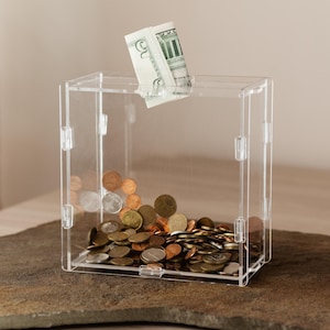 May include: A clear acrylic donation box with a dollar bill inserted into the slot. The box is filled with a variety of coins, including pennies, nickels, dimes, and quarters.