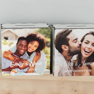 May include: Two wooden money boxes with slots on top. Each box features a photo of a couple on the front. The box on the left has a photo of a couple embracing and the text "Emily & Noah Travel Fund". The box on the right has a photo of a couple kissing and the text "Sofia & James Honeymoon Fund".