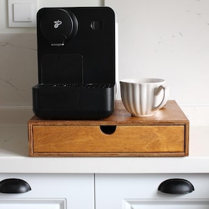 May include: A black coffee machine with a white ceramic mug on a wooden drawer. The drawer is set on a white countertop. The coffee machine has a black base and a black top with a silver logo. The drawer has a small, curved handle.