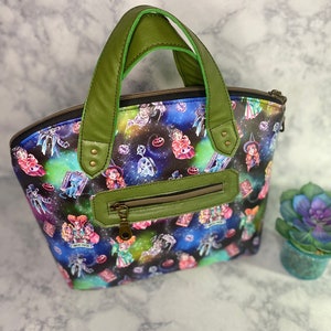 May include: A green and black tote bag with a galaxy print featuring witches and Halloween imagery. The bag has a zipper closure and two green handles.