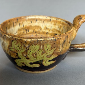 May include: A ceramic mug featuring a speckled tan and cream interior with a dark brown base. The exterior is decorated with a light green leaf-like pattern. The mug has a thick, curved handle and a wide, rounded bowl shape with a smooth finish.