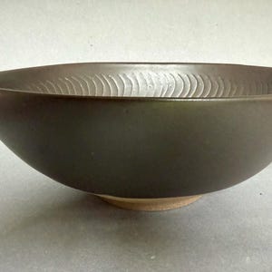 Può includere: Una ciotola in ceramica marrone scuro con esterno liscio e forma ampia e aperta. Il bordo interno presenta una serie di scanalature verticali intagliate e ripetitive che creano un motivo strutturato lungo la parte superiore.