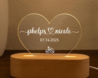 May include: A heart-shaped, illuminated acrylic sign with the names "phelps & nicole" and the date "07.14.2025" engraved. The sign sits on a wooden base, with a glowing light. A wedding or anniversary gift.