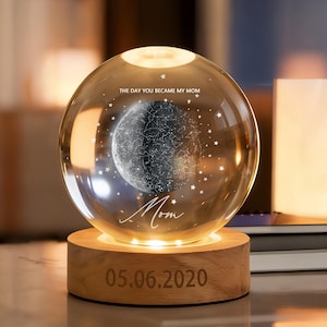 May include: A clear glass sphere with a detailed moon design and embedded stars, illuminated by a warm light. The sphere sits on a wooden base engraved with the date 05.06.2020 and the word "Mom".