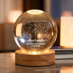 Puede incluir: Una luz nocturna de esfera de cristal transparente sobre una base circular de madera. La esfera presenta un mapa de constelaciones grabado, los nombres Oliver y Amelia, y el texto When the stars aligned 13.08.2026. La base de madera dice FOREVER & ALWAYS.