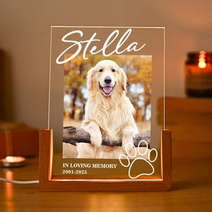 Könnte beinhalten: Eine rechteckige, klare Acrylplatte in einem hellen Holzsockel mit integrierter Beleuchtung. Die Platte zeigt ein aufgedrucktes Foto eines Golden Retrievers, den Namen Stella, den Text IN LOVING MEMORY, 2001-2025 und einen Pfotenabdruck.