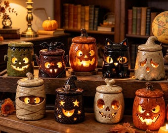 May include: A collection of nine ceramic Halloween-themed jars with lids, each featuring cut-out facial expressions illuminated from within. The designs include a green monster, a bat, a pumpkin, a black cat, a ghost, a mummy, a witch, a skull, and a jack-o'-lantern.