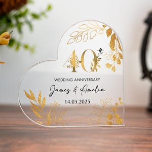 May include: Clear acrylic heart-shaped wedding anniversary keepsake. Features gold foil floral accents and the text "40th Wedding Anniversary" with names and a date. A decorative item for a special occasion.
