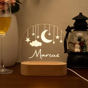 May include: A wooden-based night light with a clear acrylic panel featuring a crescent moon, stars, and a cloud design. The name "Marcus" is written in a script font. The light is plugged in with a white cord.