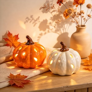 May include: Two ceramic pumpkin-shaped lanterns, one orange and one white, each featuring a brown stem and cut-out openings that emit a warm internal glow. The pumpkins are arranged with scattered autumn leaves and a vase of dried flowers.
