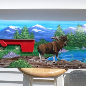 May include: Hand-painted mailbox featuring a moose standing near a lake with mountains in the background. The mailbox has a red flag and is painted with shades of blue, green, and brown. The mailbox is mounted on a wooden stand.
