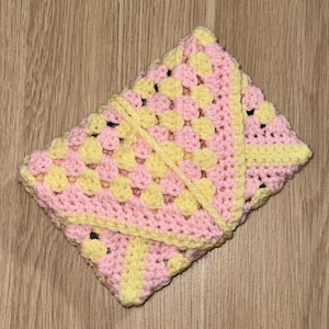 May include: A rectangular crochet pouch featuring a granny square pattern in alternating pastel pink and pale yellow yarn. The pouch has a triangular flap closure secured by a matching crocheted cord that wraps around the exterior of the item.