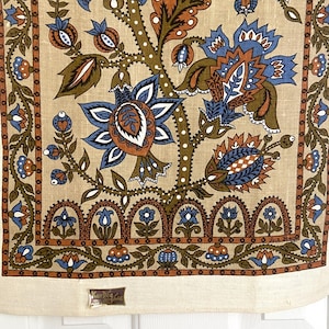 May include: A beige linen tea towel with a floral pattern in shades of blue, brown, and orange. The pattern features a large central flower with smaller flowers and leaves surrounding it. The towel has a border of brown and blue geometric shapes.